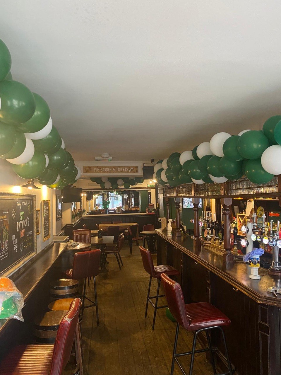 The bar inside The Cricketers Arms decorated with green and white balloon garlands along the ceiling for St Patrick's Day, with hand pumps and leather bar stools along the counter