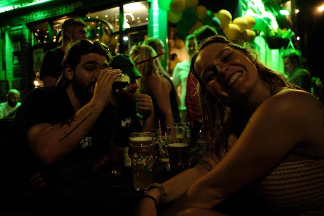 Two customers smiling over pints at an outdoor table outside The Cricketers Arms during an evening St Patrick's event, lit green, with balloons and a crowd behind them