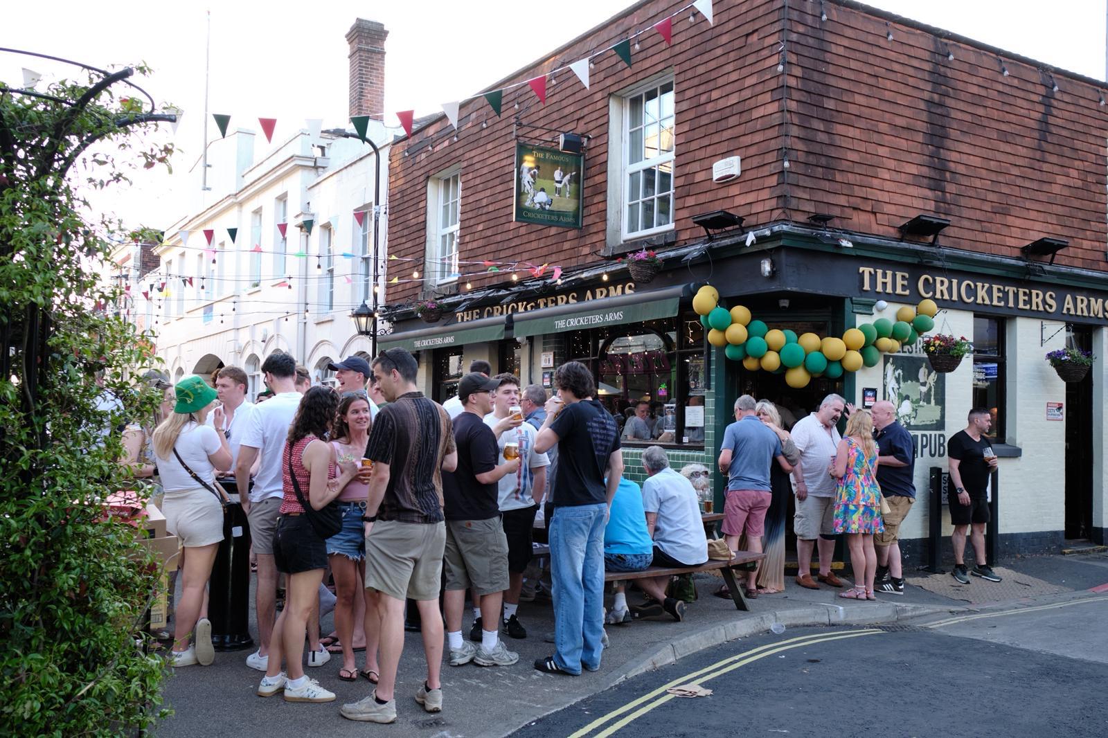 A busy sunny day outside The Cricketers Arms, with a crowd drinking at the tables and on the pavement under bunting and a green and gold balloon arch