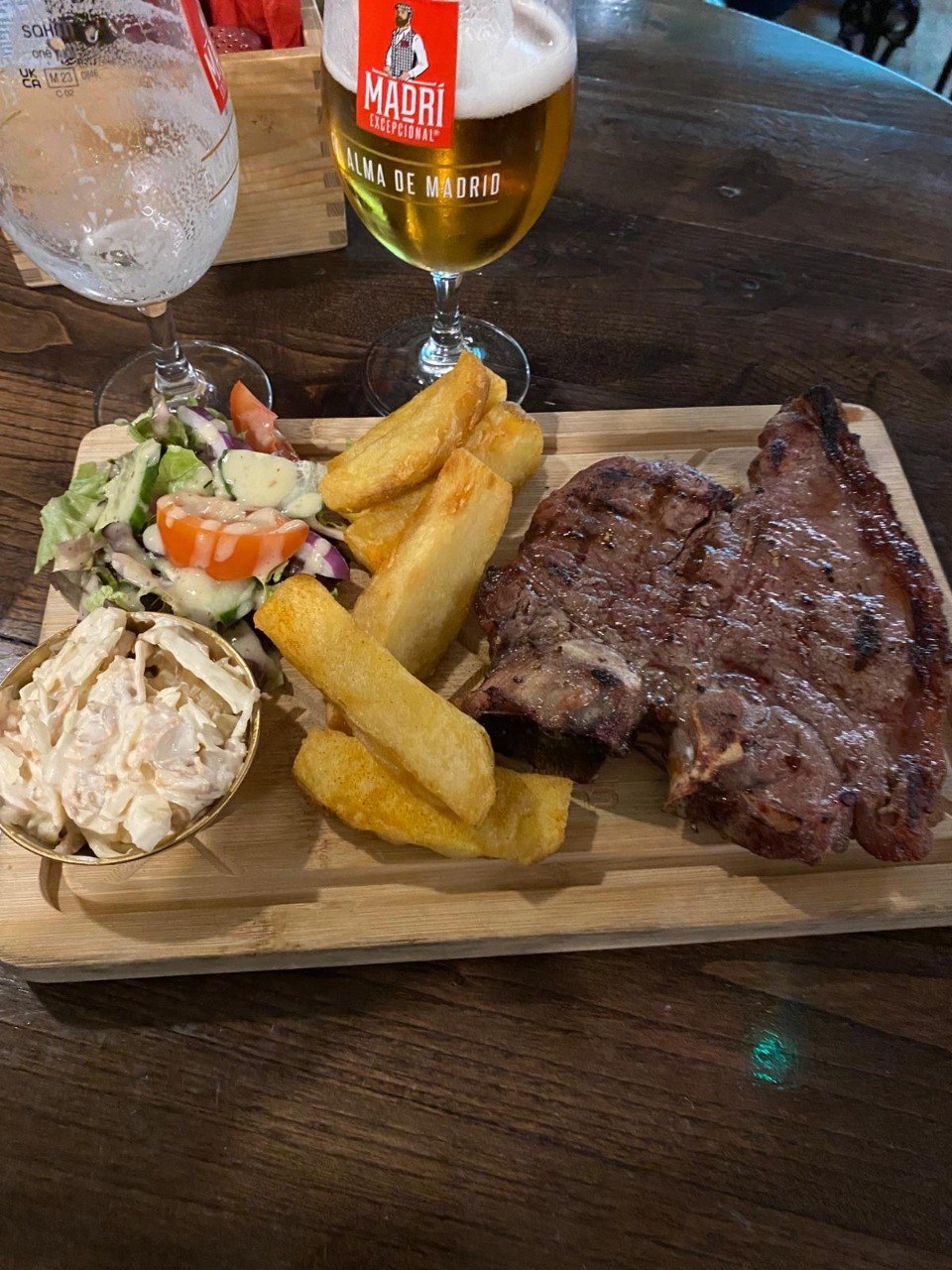 A steak on a wooden board with chunky chips, salad and coleslaw, served with a beer and a glass of wine