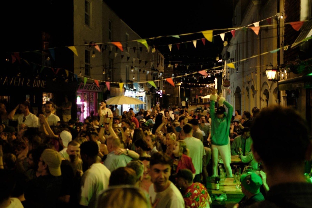 A large crowd packing the street outside The Cricketers Arms at night, with colourful bunting strung overhead and one person standing on a table with their arms raised