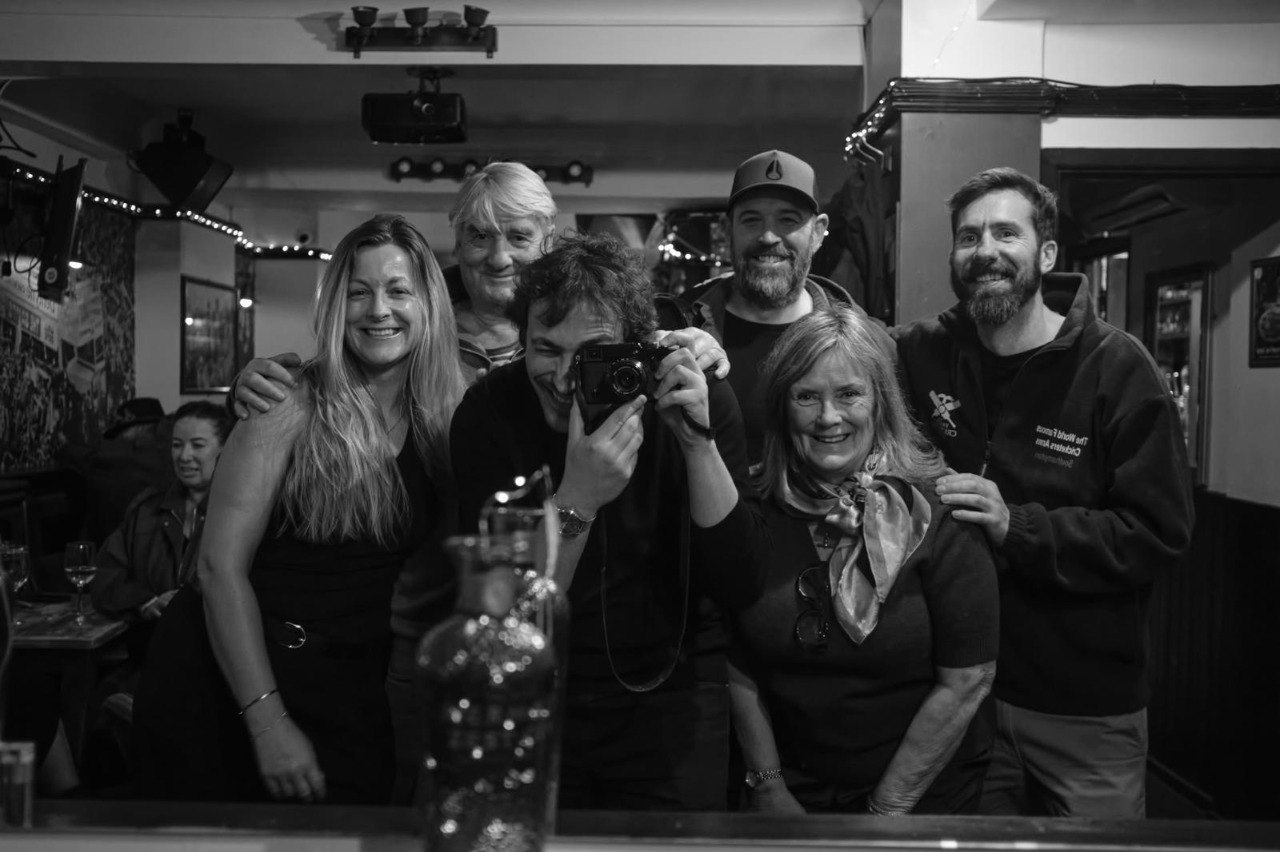 A black and white group photograph of six people posing together inside The Cricketers Arms, one holding a camera and one wearing a World Famous Cricketers Arms sweatshirt