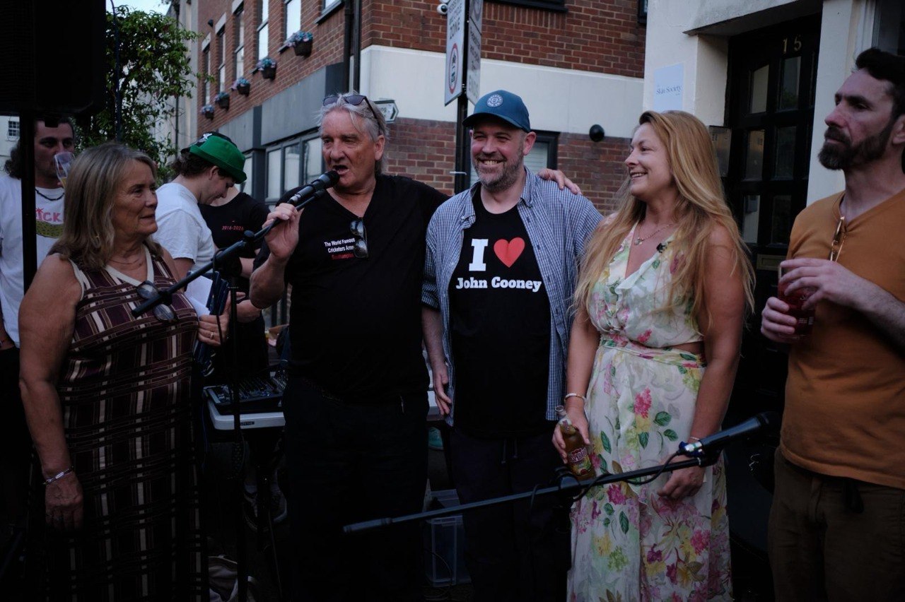 The landlord speaking into a microphone outside The Cricketers Arms at a street event, with members of the team and regulars gathered alongside him