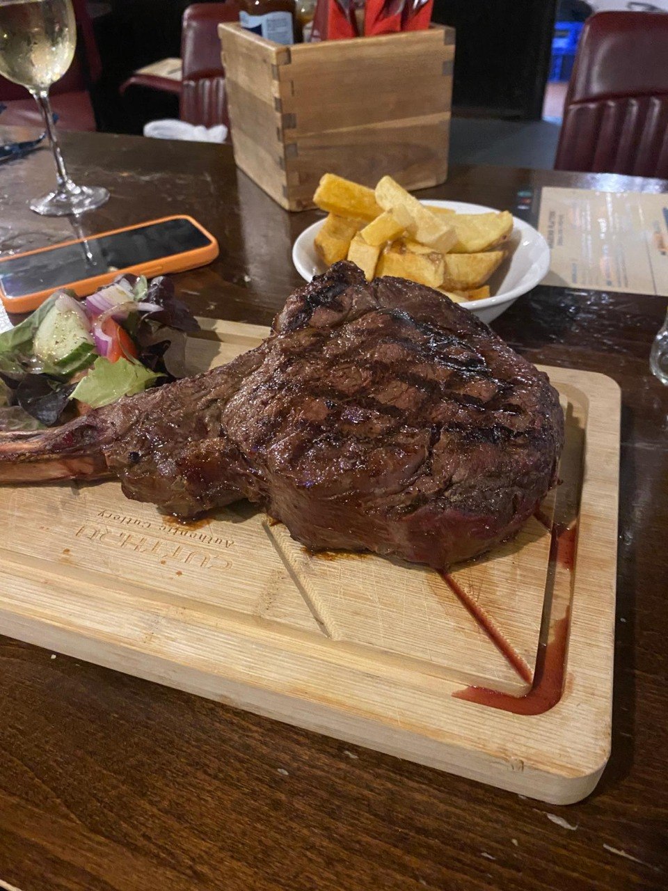 A chargrilled tomahawk steak on a wooden serving board with a bowl of chunky chips and a side salad